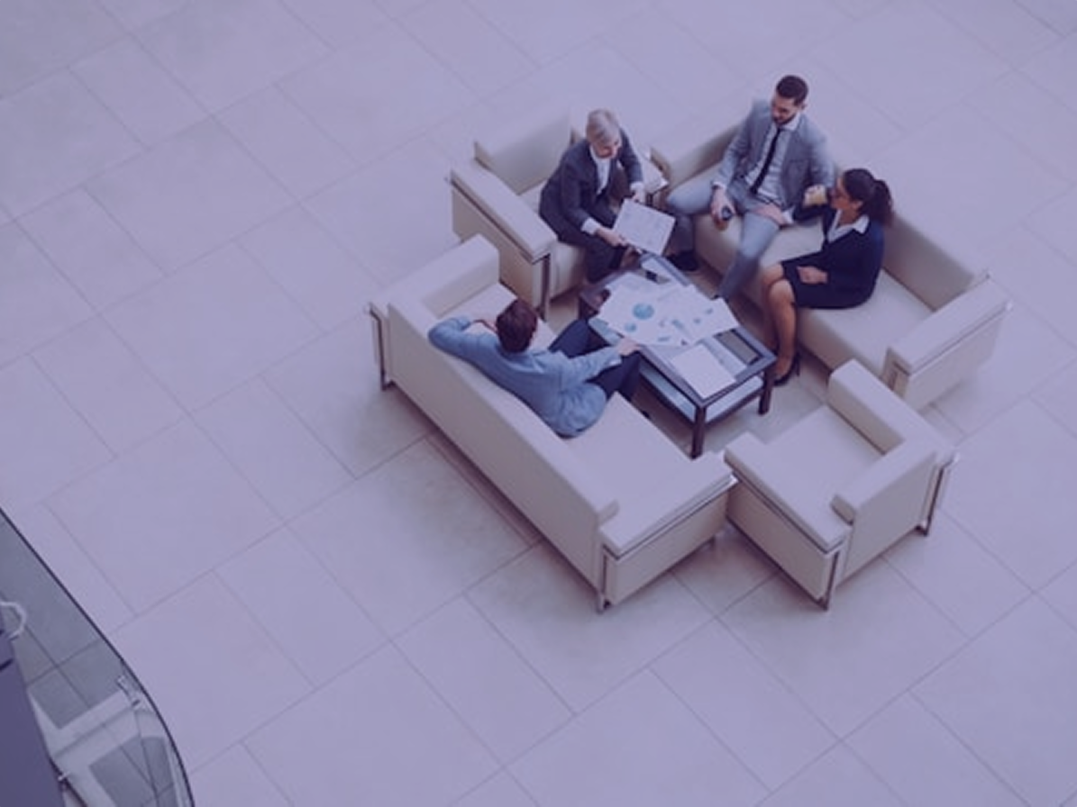 Overhead view of four colleagues meeting on couches in an office lobby, charts spread across the coffee table